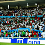**

"Bustling scene at the Muscat International Stadium during a football (soccer) World Cup qualifier match, fully clothed athletes in action, enthusiastic crowd in the stands, flags waving, safe for work, appropriate content, professional sports photography, perfect anatomy, natural proportions, family-friendly atmosphere, modern stadium architecture."

**